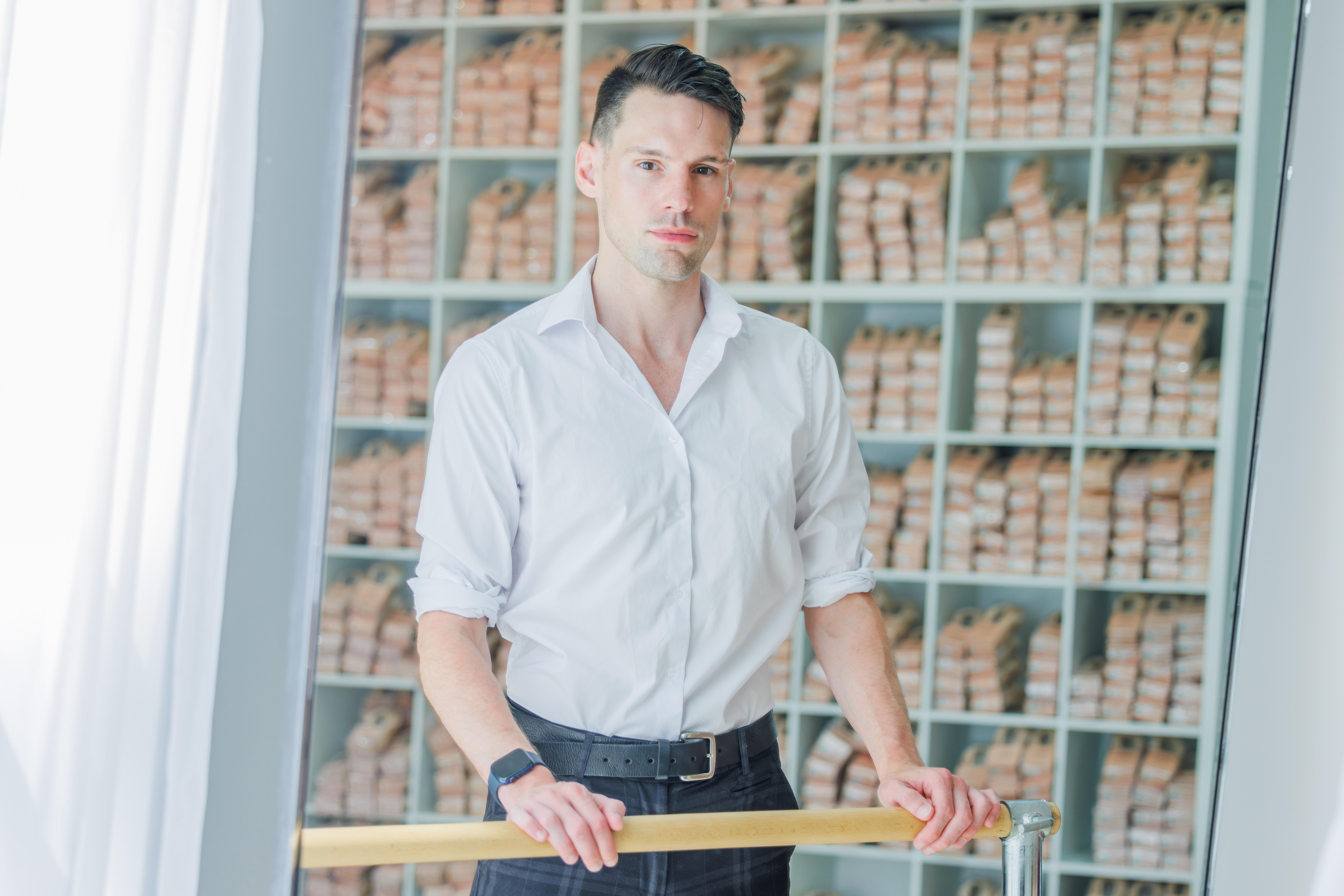 Riley Weber stands at ballet barre with rows of pointe shoes behind him